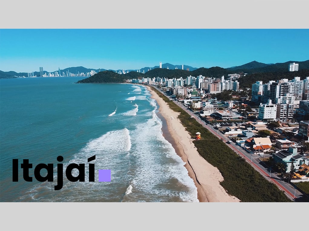 Vista aérea da praia de Itajaí, com ondas e prédios ao fundo. Itajaí em destaque.