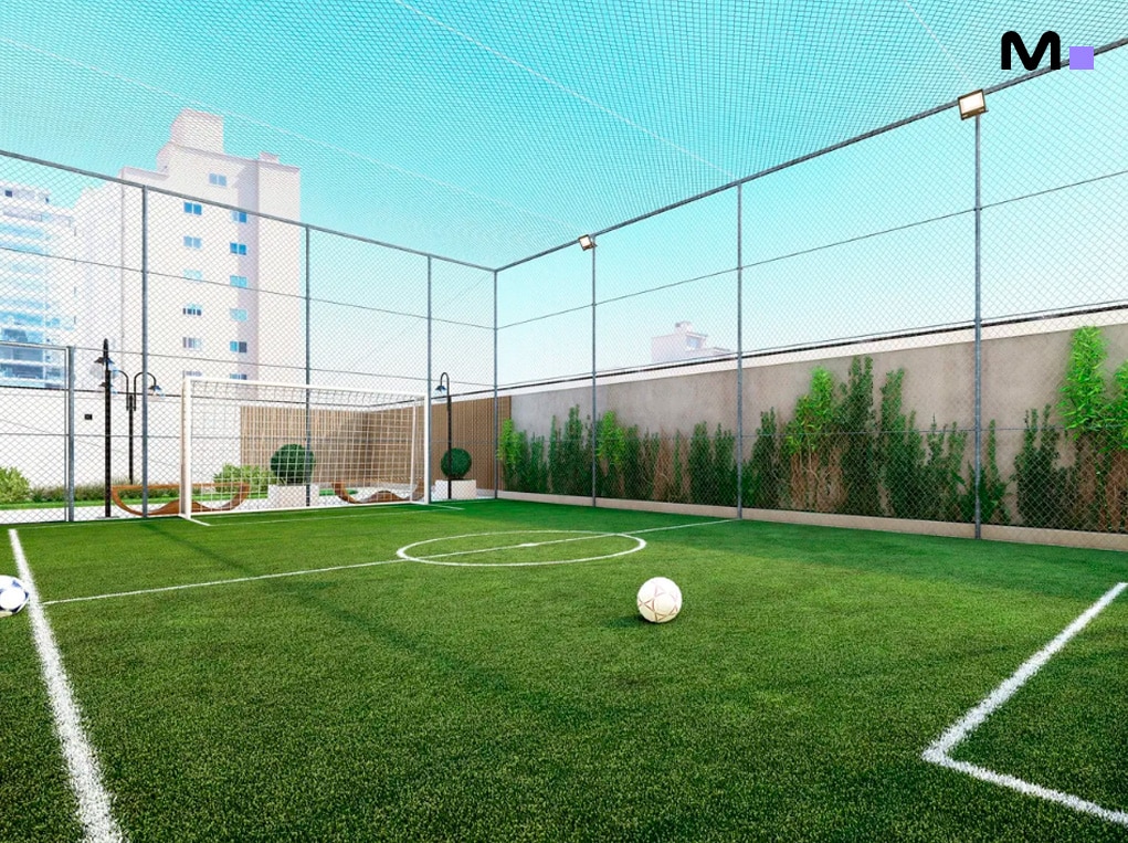 Campo de futebol society com grama sintética, cercado por rede e prédios ao fundo.