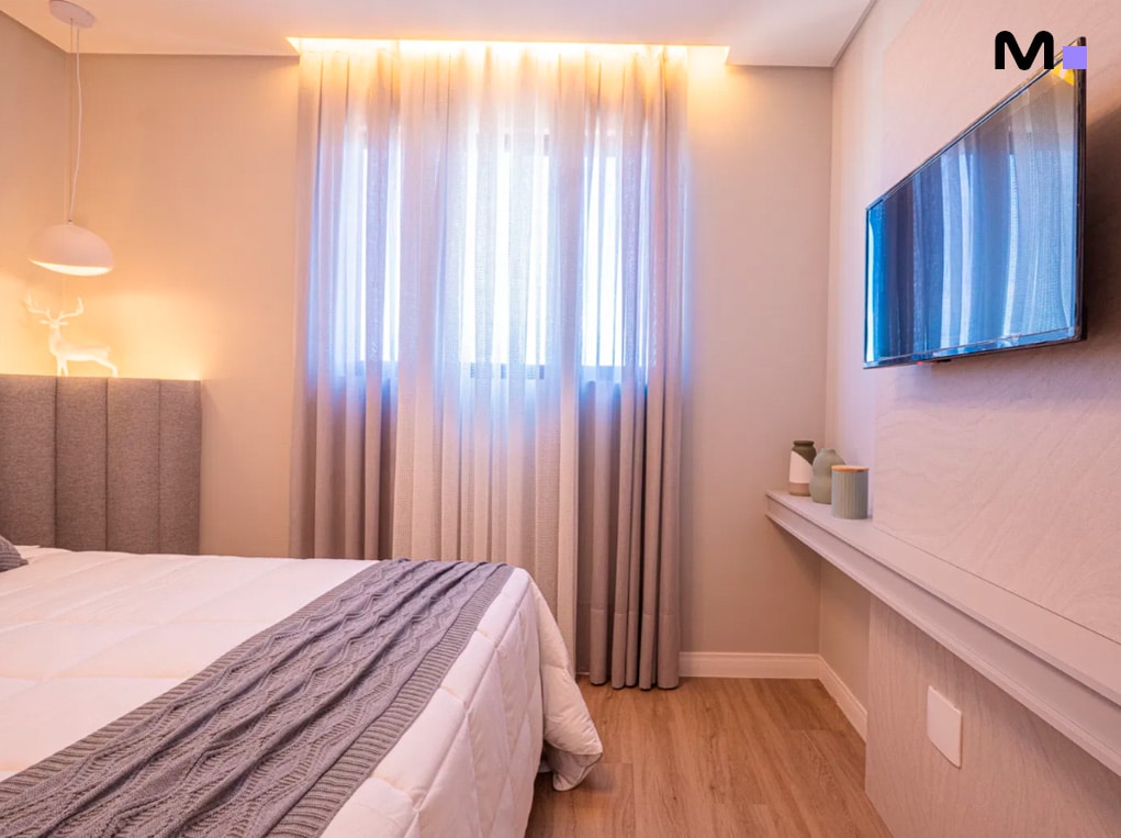 Quarto moderno com cama, TV e cortinas luminosas. Edifício Leblon Residence.