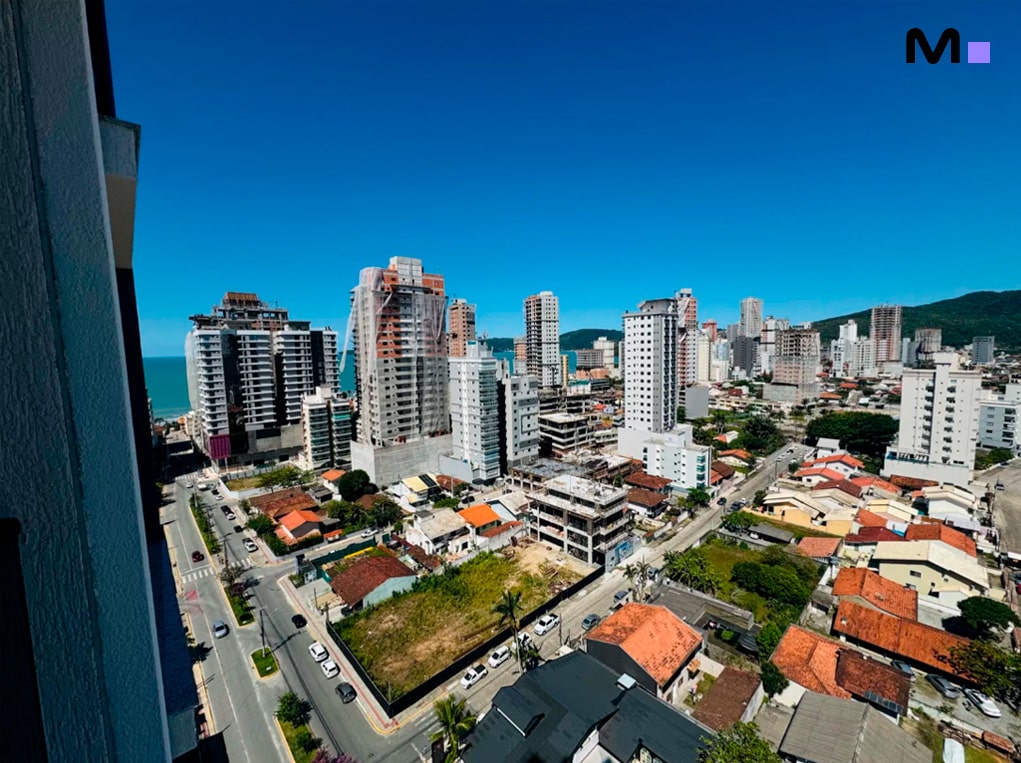 Vista aérea de Balneário Camboriú com o Edifício Ocean Cay ao fundo. Céu azul e prédios modernos.