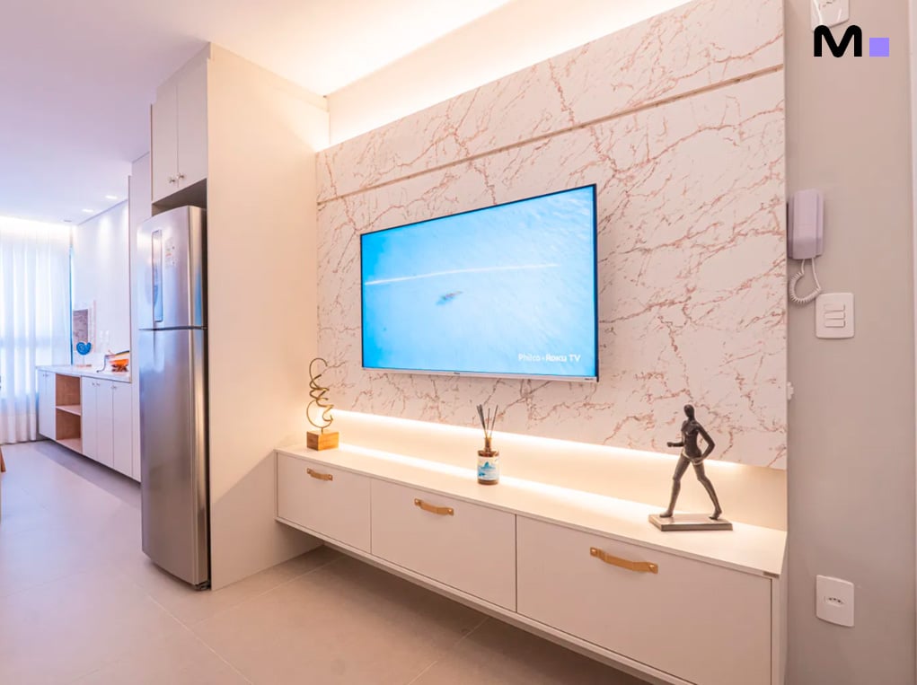 Sala moderna com TV e painel decorativo no Edifício Leblon Residence. Armários brancos e iluminação suave.