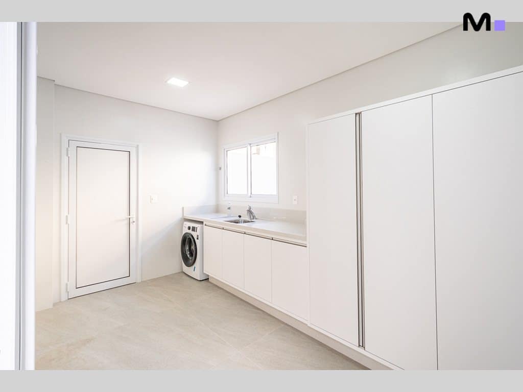 Lavanderia branca e minimalista no Condomínio House Haras com máquina de lavar e armários.