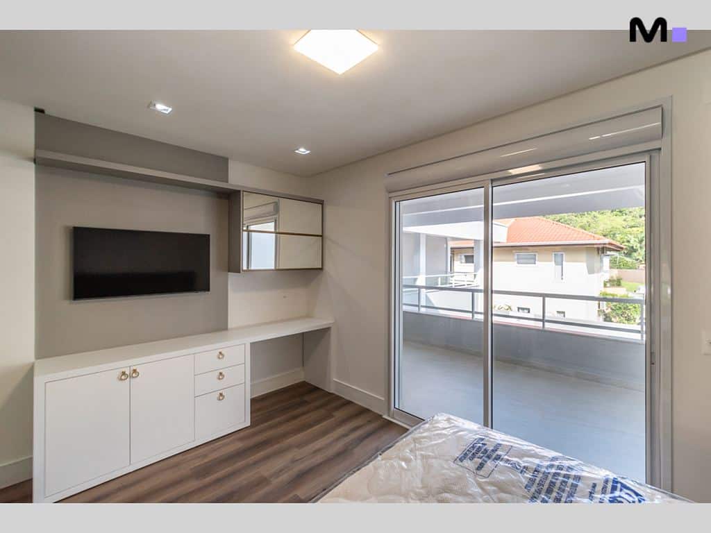 Quarto moderno no Condomínio House Haras com TV, armários brancos e varanda com vista.