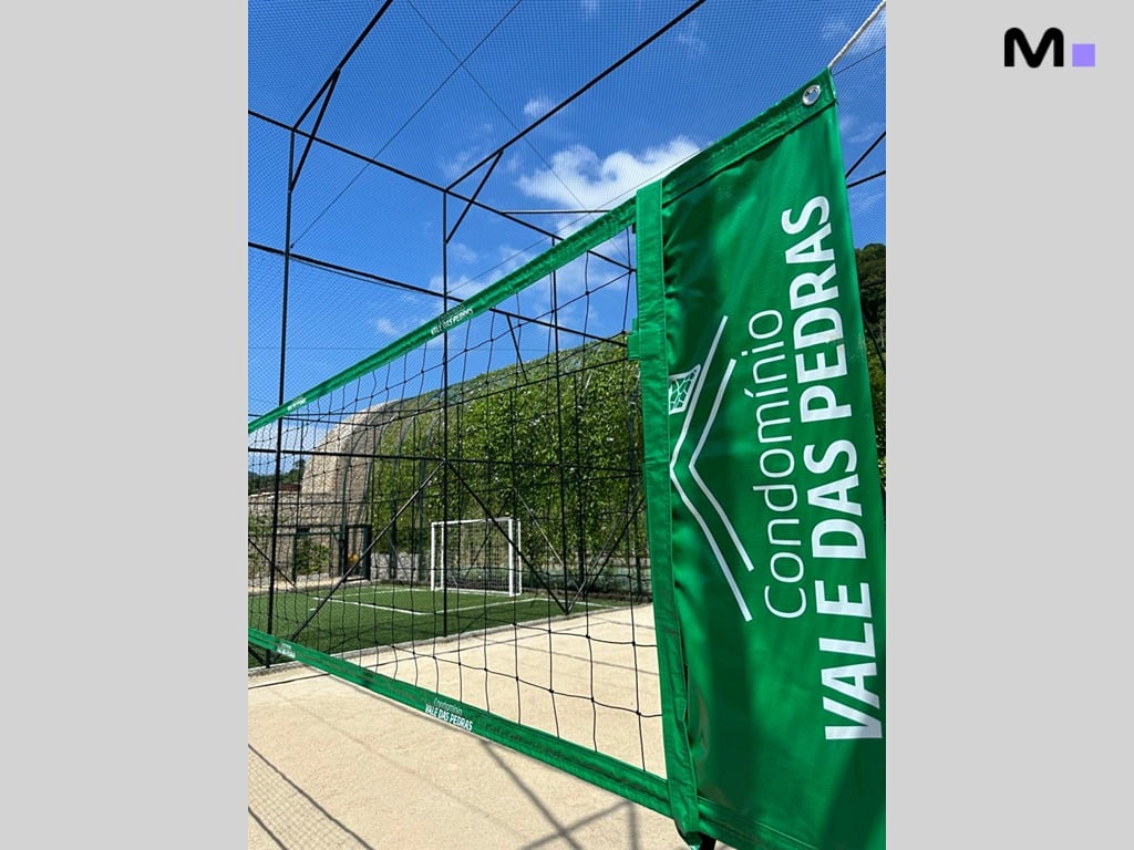 Quadra esportiva do Condomínio Vale das Pedras com rede de vôlei e campo de futebol ao fundo.