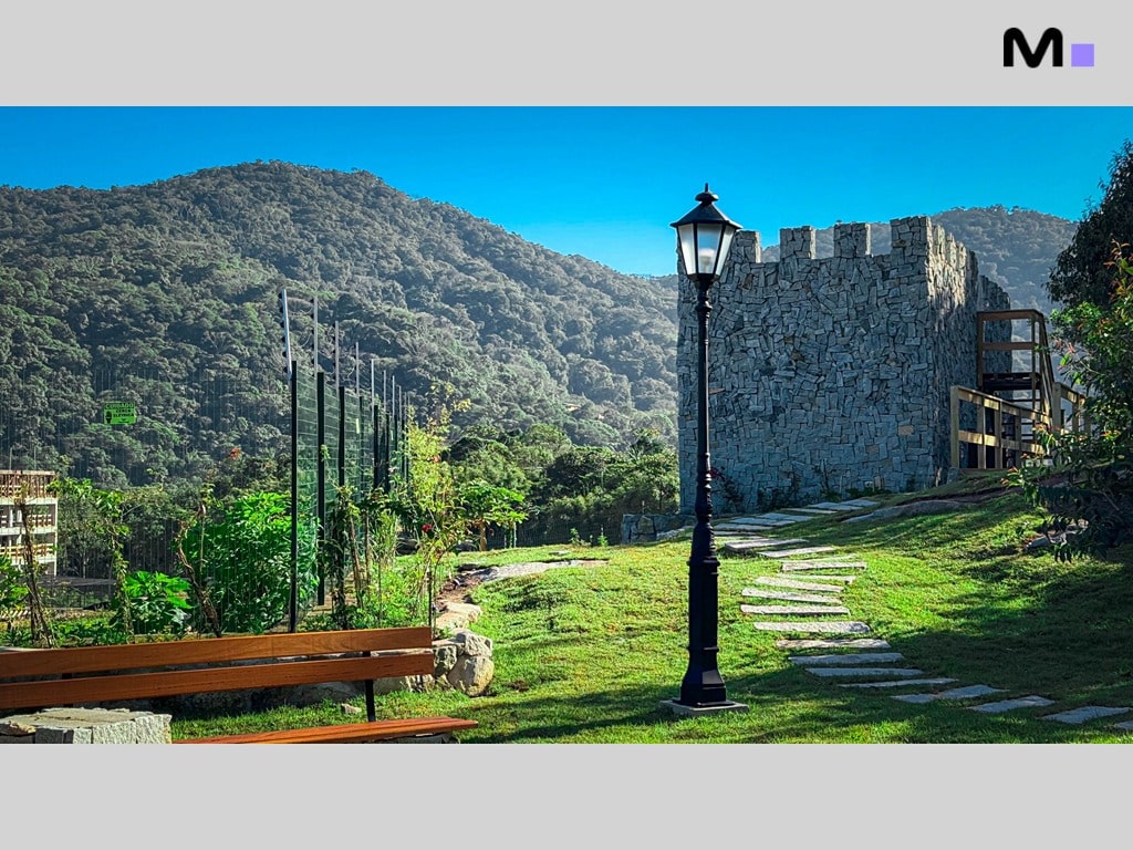 Paisagem do Condomínio Vale das Pedras com torre de pedra, banco e montanhas ao fundo.