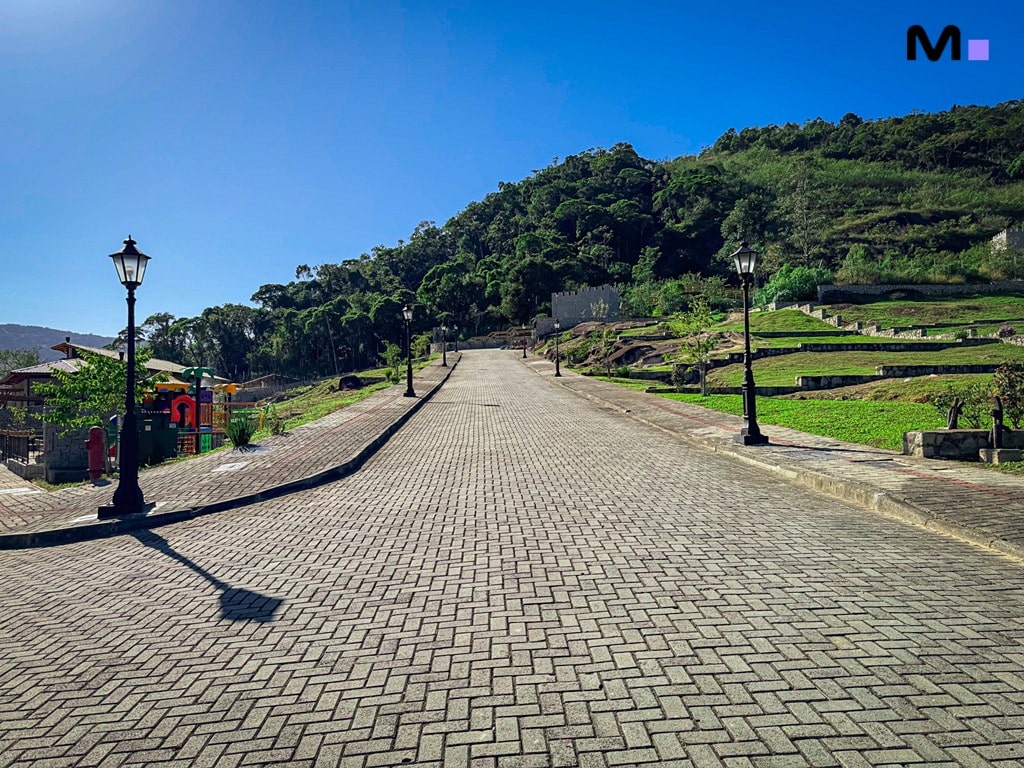 Rua de paralelepípedos no Condomínio Vale das Pedras, com iluminação e paisagem verde. Céu azul.