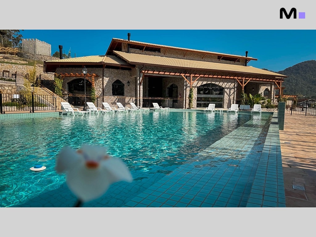 Piscina azul do Condomínio Vale das Pedras com casa de pedra ao fundo.