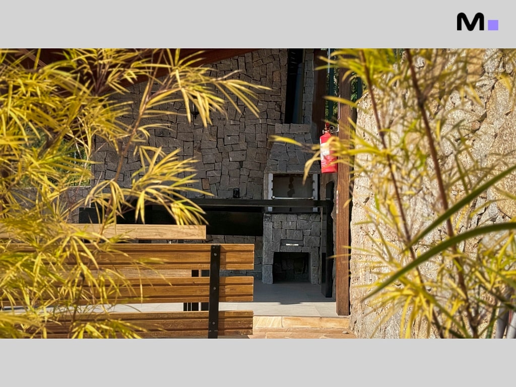 Churrasqueira de pedra no Condomínio Vale das Pedras, com detalhes em madeira e vegetação.