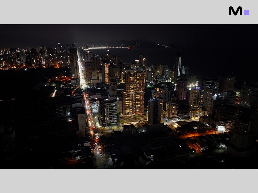 Visão noturna iluminada de Balneário Camboriú, com arranha-céus e luzes da cidade refletindo no oceano.